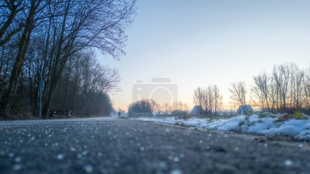 Ten niskokątny ujęcie oddaje istotę ostry zimowy poranek na wiejskiej drodze. Pierwsze światło świtu oświetla niebo, rzucając delikatny blask nad sceną. Droga jest nakrapiana