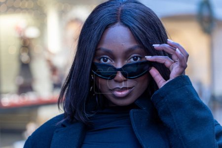 This image captures a young African American woman posing with style on an urban street. Shes lowering her sleek black sunglasses with one hand, revealing a confident gaze. The blurred background