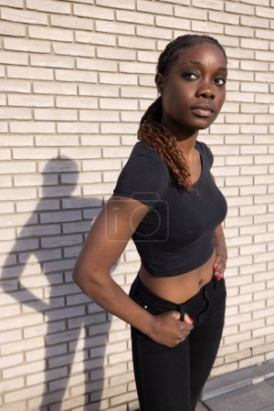 This image captures a young African woman confidently posing in front of a white brick wall. The sunlight casts her shadow prominently beside her, creating a dramatic effect. She wears a casual black