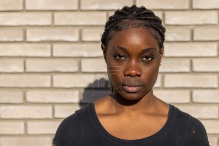 This image showcases a young African woman standing confidently in front of a white brick wall. The subject is centered in the frame, with her hair neatly styled in cornrows that add a textural