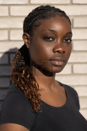 This compelling image features a close-up of a young African woman with a confident and direct gaze. The natural daylight accentuates her facial features and the intricate braids in her hair, which