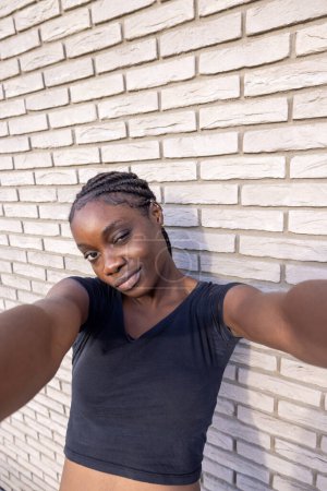 Téléchargez les photos : Il s'agit d'un selfie intime d'une jeune femme africaine aux cheveux tressés, avec un sourire ludique alors qu'elle tend la main vers la caméra. Elle porte un haut noir décontracté et se positionne contre un - en image libre de droit