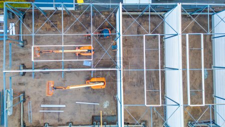 Cette image aérienne offre une vue aérienne d'un chantier de construction, mettant en valeur l'agencement géométrique des échafaudages et des machines. Deux ascenseurs télescopiques orange se détachent contre la boue