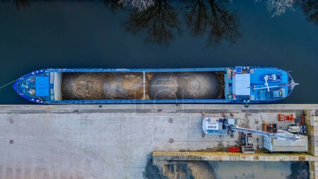 The image presents a top-down aerial view of a blue cargo ship docked at a concrete quay. The ship is being loaded or unloaded with materials, evidenced by the open hatch compartments filled with
