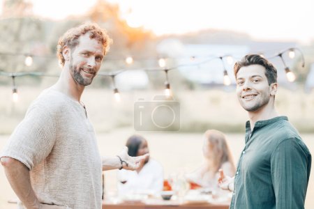 Esta fotografía captura a dos hombres en una reunión al aire libre durante lo que parece ser una noche de verano. El hombre en primer plano está vestido con una camisa de lino casual y tiene el pelo rizado, presentando una