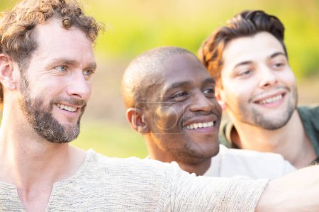 This image portrays a group of three men of different ethnicities sharing a joyful moment outdoors. The man in the foreground, with curly hair and a light stubble, is looking away with a smile, as if