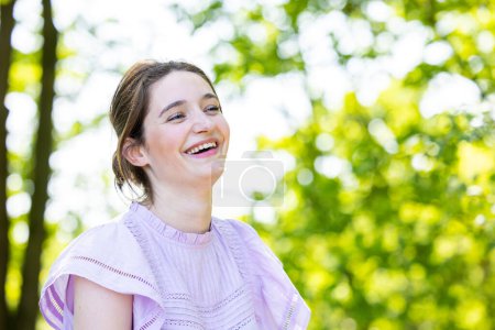 This image features a young woman outdoors, bathed in natural light, with a backdrop of vibrant green foliage indicative of a summer park. She is dressed in a delicate, lilac-colored dress with cut