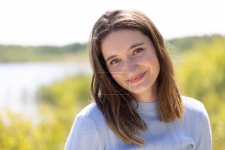 This image features a young woman with a soft, serene smile, standing by a lakeside. She is dressed in a light blue sweater, which adds a gentle touch to the overall warmth of the scene. The