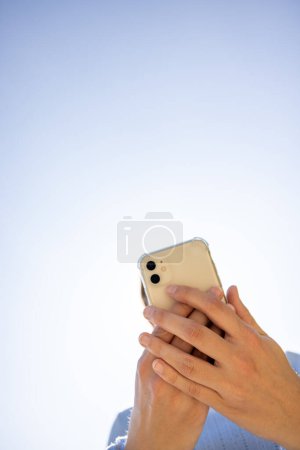 Téléchargez les photos : Cette image montre une personne mains tenant un smartphone sur un fond de ciel clair. L'angle de prise de vue est d'en bas, en se concentrant sur l'appareil et les mains qui interagissent avec lui. La simplicité de - en image libre de droit