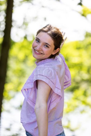 This image shows a young woman turning to smile over her shoulder, her eyes gleaming with playfulness and joy. She is outside on a sunny day, with trees providing a dappled light backdrop. Her light