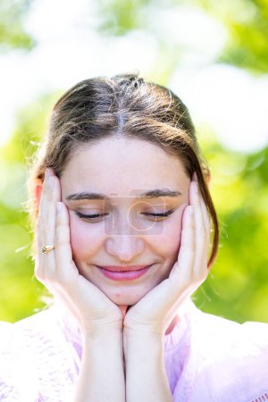 Photo for This portrait showcases a young woman gently holding her face in her hands, her eyes closed and a soft smile playing on her lips. The image exudes a sense of peace and self-reflection. The blurred - Royalty Free Image