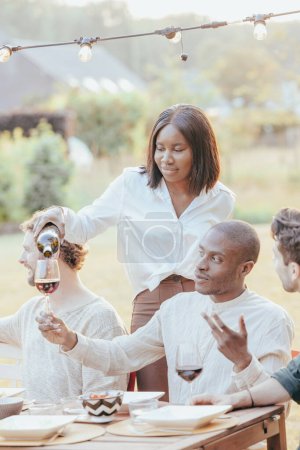 The photograph depicts a black african american woman attentively pouring wine into a guests glass during an outdoor multi racial dinner party. The scene is set in a warm, glowing atmosphere as the