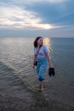 Das Bild zeigt eine junge Frau, die bei Sonnenuntergang gemütlich in den ruhigen Gewässern watet. Mit Schuhen in der Hand scheint sie das Gefühl des Wassers an ihren Füßen zu genießen, ihr Blick richtet sich