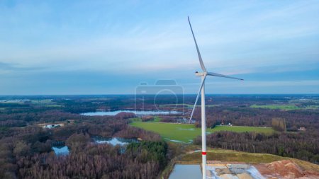This aerial image features a solitary wind turbine dominating the foreground, with its blades reaching towards a spacious sky. Below, a patchwork of forest and fields stretches into the distance