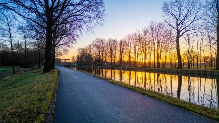 This tranquil image captures a serene sunrise along a still canal, flanked by a path and a row of tall, bare trees. The rising sun infuses the sky with shades of orange and yellow, casting reflections