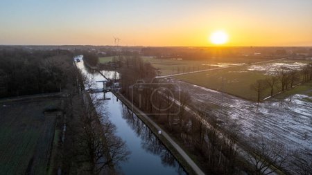 Foto de Esta fotografía aérea captura un crujiente amanecer de invierno, con el sol flotando justo por encima del horizonte, arrojando una suave luz dorada a través del paisaje. El canal que atraviesa la escena refleja el - Imagen libre de derechos