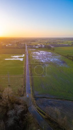 This aerial image captures the tranquil beauty of the countryside during sunset. A straight country road cuts through the landscape, leading towards the horizon where the suns final glow graces the
