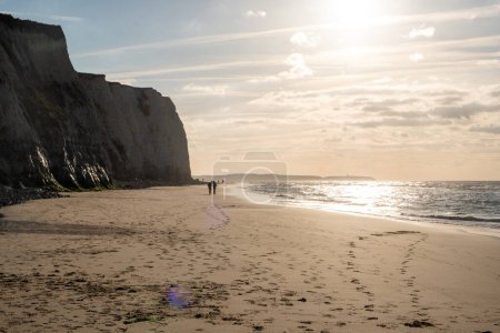 Photo for This image is characterized by the tranquil ambiance of an evening walk on a beach flanked by towering cliffs. The setting sun casts a soft, diffused light that highlights the texture of the cliff - Royalty Free Image