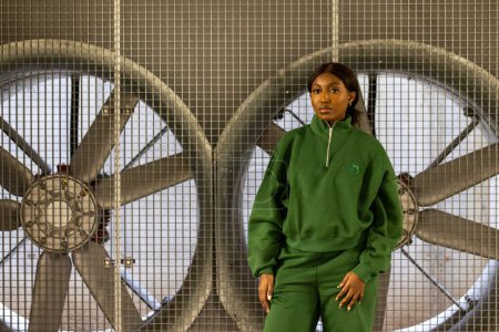 In this industrial setting, an African American woman stands confidently in a casual green tracksuit. Behind her, two large metallic fans enclosed in safety cages create a symmetrical, visually