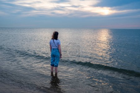 The image features a person in a moment of solitude, standing in the shallow waters of the sea as dusk settles in. The individual is dressed in rolled-up jeans and a casual shirt, suggesting a relaxed