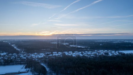 An aerial perspective captures the serene beauty of a winter morning as the first light of dawn breaks across a snow-dusted countryside. Contrails in the sky suggest the early stirrings of distant