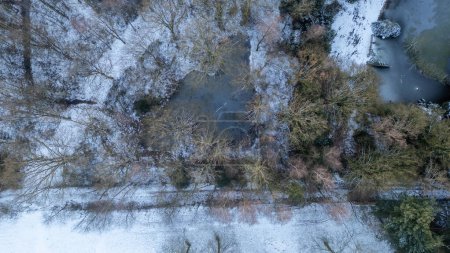 This aerial image reveals a serene winter landscape where a narrow waterway meanders through a frosted terrain. The trees, dusted with snow, create delicate patterns that weave through the ground