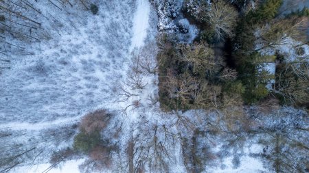 Widok z powietrza oddaje spokojny zimowy krajobraz, gdzie kurz śniegu pokrywa ziemię, tworząc delikatną mozaikę z odkrytą ziemią. Kompozycja jest zrównoważona drogą nawijania lub