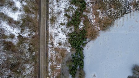 Dieses Foto bietet einen Blick aus der Vogelperspektive auf eine einsame Straße, die durch eine winterliche Landschaft führt. Auf der einen Seite ein Flickenteppich aus schneebedeckten Feldern und kahlen Bäumen, auf der anderen eine dichte Ansammlung von