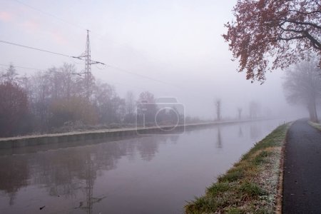 Téléchargez les photos : Cette photographie capture une scène tranquille tôt le matin par un canal, voilé dans un brouillard dense et doux. La brume enveloppe les arbres environnants et les lignes électriques, créant un paysage minimaliste qui transmet - en image libre de droit