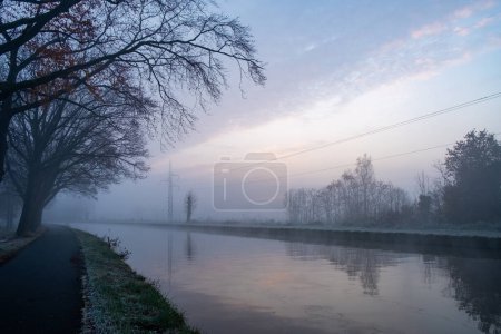A hauntingly beautiful dawn breaks over a canal, the scene shrouded in a thick mist that diffuses the morning light. The silhouette of leafless branches reaches out over the water, where the first