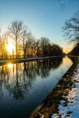 This vibrant image captures the dynamic contrast of a bright winter sun setting behind bare trees, reflecting its brilliance on the still waters of a canal. The sun, positioned just at the edge of the