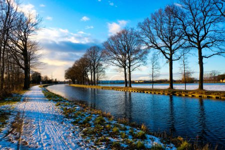 Dieses fesselnde Bild zeigt einen schneebedeckten Pfad, der entlang eines ruhigen Kanals verläuft, flankiert von majestätischen, blattlosen Bäumen vor einem strahlenden Winterhimmel. Die untergehende Sonne wirft ein sanftes Licht