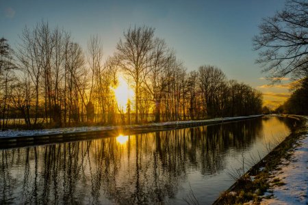 This image depicts a stunning sunset scene where the suns golden rays pierce through the silhouetted trees along a calm canal. The sky, ablaze with warm tones, transitions from yellow to a deep blue