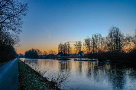 Téléchargez les photos : La photographie présente un moment serein avant l'aube sur un canal partiellement gelé. La lumière du matin pénètre doucement dans le ciel, peignant un gradient doux du blues frais aux oranges chaudes à la - en image libre de droit