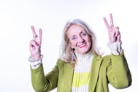 The image captures an elderly woman with a vivacious spirit, her face lit up with a youthful smile as she makes the peace sign with both hands. Her attire, a chic lime green blazer paired with a