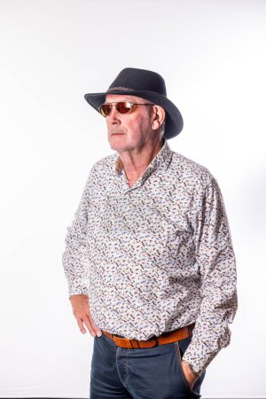This image presents a senior man looking off into the distance, wearing a casual patterned shirt and a stylish fedora hat paired with sunglasses. The relaxed fit of his shirt and the comfortable