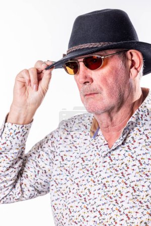 Dieses Bild zeigt einen älteren Mann, der leicht seine Sonnenbrille aufsetzt und einen geheimnisvollen und faszinierenden Blick bietet. Sein ernster Gesichtsausdruck und die Einstellung seiner Sonnenbrille deuten auf einen Moment der Ruhe hin.