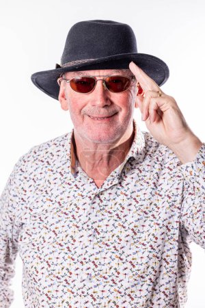 This engaging image features a senior man with a confident smile, tipping his fedora hat in a greeting or acknowledgment. His stylish sunglasses add an air of coolness and mystery. The mans playful