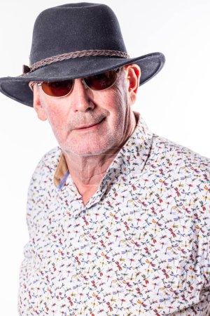 Dieses Porträt zeigt einen älteren Mann, der Zuversicht und Stil ausstrahlt. Er trägt einen dunklen Fedora-Hut gepaart mit einer klassischen Sonnenbrille, ein Look, der Aufmerksamkeit verdient. Sein direkter Blick in die Kamera