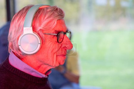 This image features a profile view of an elderly man gazing out of a window, lost in thought. He wears a pair of modern, over-ear headphones and a burgundy sweater, which suggests a blend of