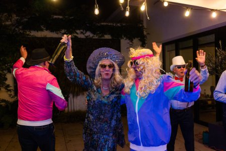 The image captures a group of people, dressed in retro fashion, enjoying a lively dance at a themed party. A person in the center, adorned with a sparkling hat and sunglasses, is raising a bottle