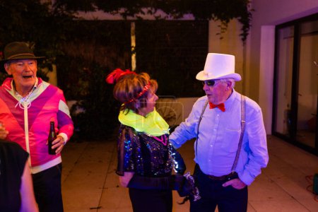 The image showcases a lively moment among senior friends at a casual evening gathering. A man in a white cowboy hat and bow tie engages in conversation with a woman adorned with a colorful scarf, her