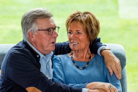 The image depicts an endearing moment between a senior couple as they sit closely together on a comfortable couch. The man, dressed in a smart navy cardigan over a light blue shirt, gazes fondly at