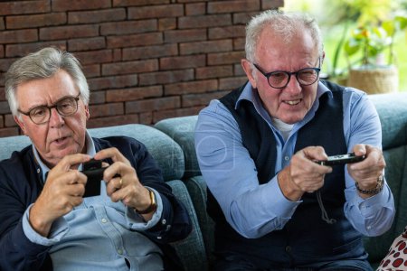 This lively image captures two senior men deeply engrossed in playing video games, their faces animated with concentration and glee. The man on the left is dressed in a smart casual attire with a blue