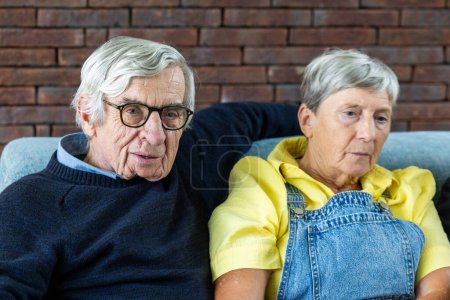 This candid image captures an elderly couple side by side, sharing a quiet moment on their comfortable sofa. The man looks towards the camera with a gentle smile, sporting glasses and a classic navy