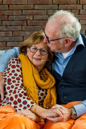 This warm photograph captures an elderly couple sharing a tender moment. The man is leaning in to affectionately whisper to his partner, who listens with a gentle and contented smile. The womans