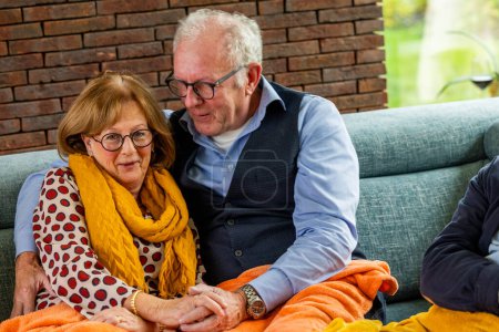 The photograph showcases an elderly couple sharing a cozy and affectionate moment on a couch. The woman, donning glasses and a warm yellow scarf, offers a gentle smile to the camera, while the man