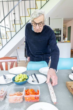 This image depicts a senior man thoughtfully preparing a meal in a bright, modern kitchen. Hes arranging plates on the table, suggesting a careful and considerate approach to mealtime. The table is
