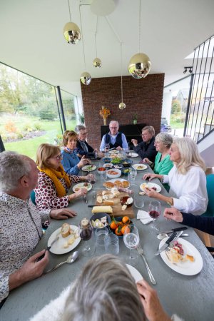 Dieses ermutigende Bild zeigt eine Gruppe Senioren, die sich um einen Esstisch versammelt haben, um in einem hellen und modernen Raum gemeinsam zu essen. Die großen Fenster bieten einen Blick auf den Garten und verbinden diese miteinander