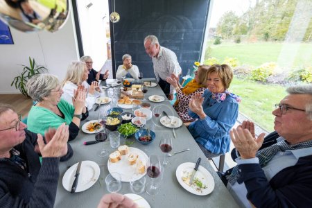 A group of seniors enjoys a convivial meal together around a dining table filled with an assortment of dishes and wine. A man stands, possibly giving a toast or sharing a story, as he is greeted with